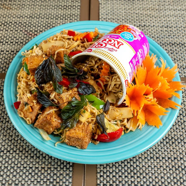 Noodles with tofu, vegetables, basil, and a Nissin Cup Noodles container on a blue plate.