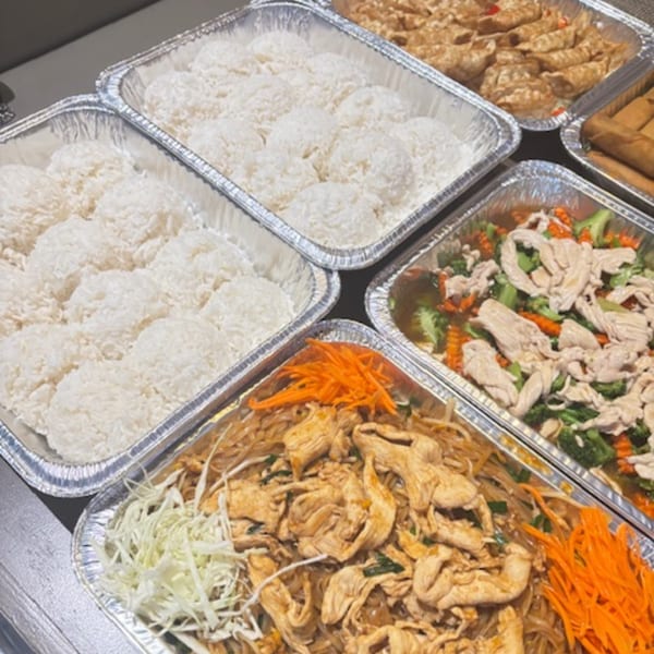 Multiple foil trays filled with Asian cuisine, including rice, noodles, chicken, and vegetables.