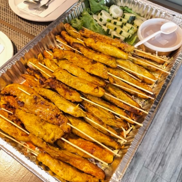 A catering tray filled with numerous chicken satay skewers, lettuce, sliced cucumbers, and a dipping sauce.