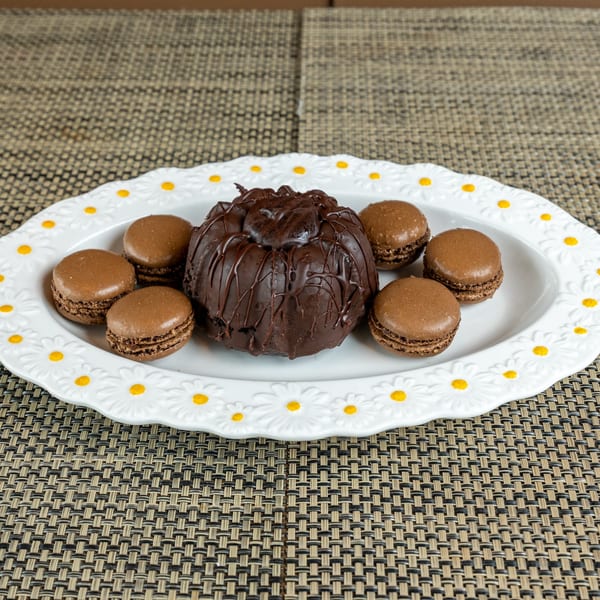 A chocolate bundt cake with chocolate drizzle and six chocolate macarons on a daisy-patterned plate.