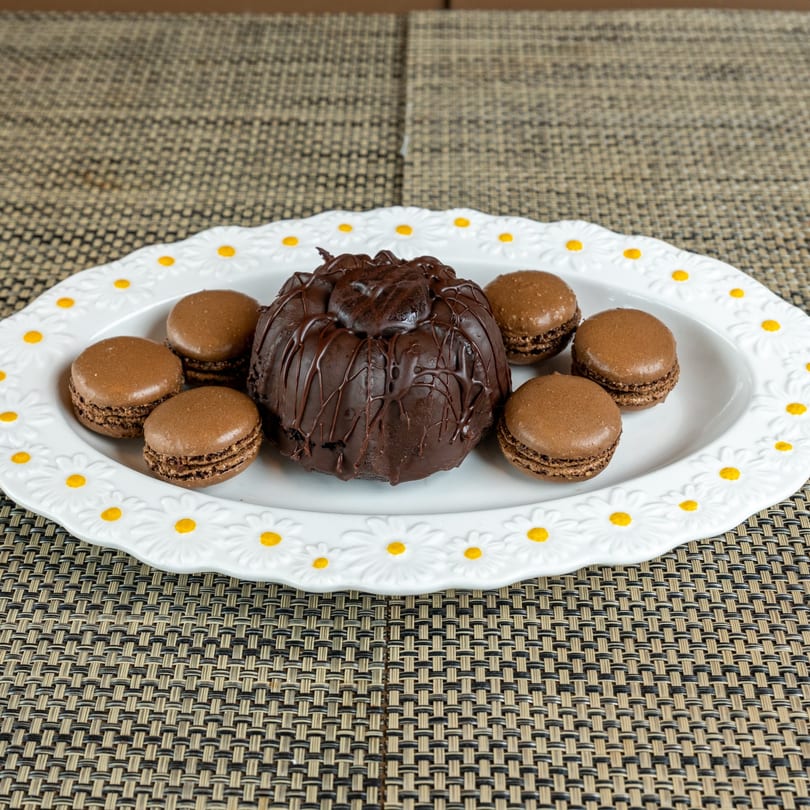 A chocolate bundt cake with chocolate drizzle and six chocolate macarons on a daisy-patterned plate.