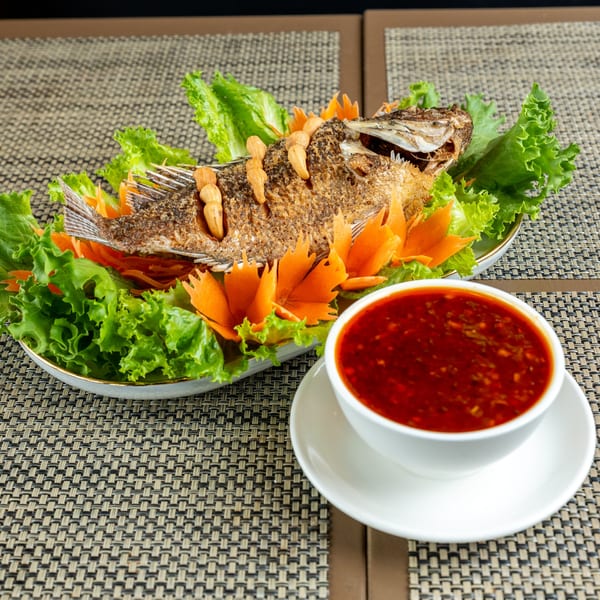Crispy fried whole fish garnished with lettuce and carrot flowers, served with a side of red chili sauce.